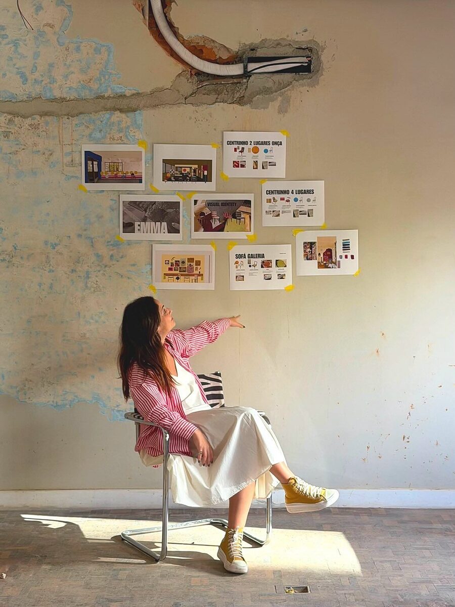 Rapha sentada apontando para os moodboards do Emma colados numa parede com pátina, com painéis de identidade visual, sofá galeria e centrinhos
