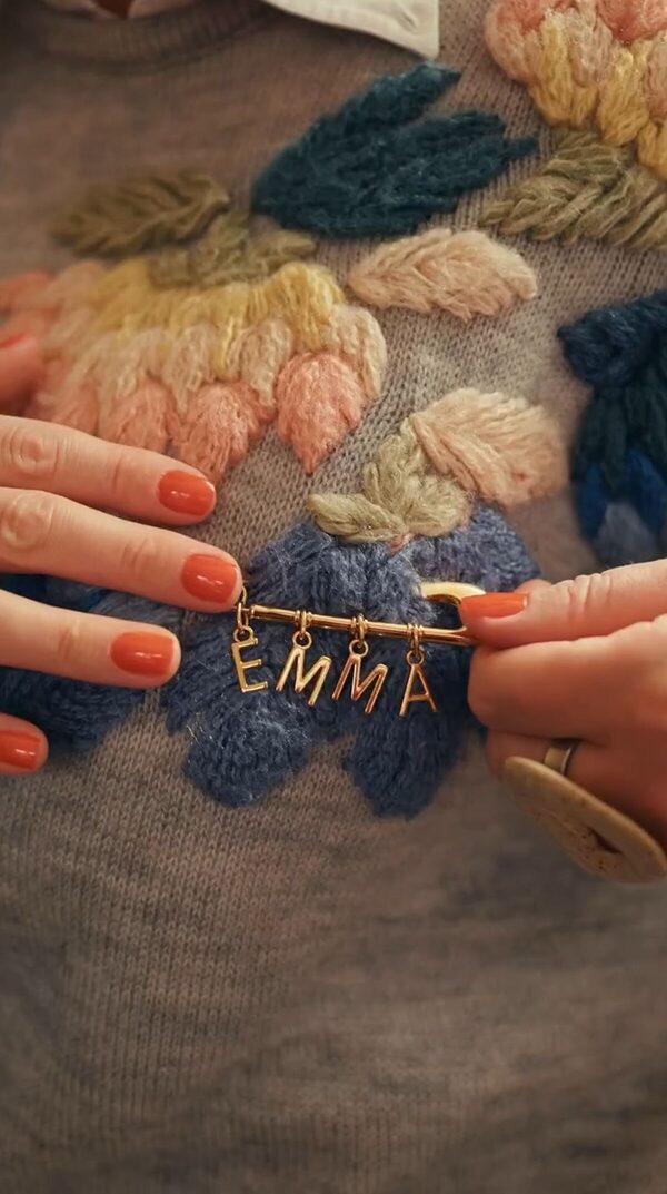 Mão segurando broche dourado com o nome Emma sobre tricô bordado