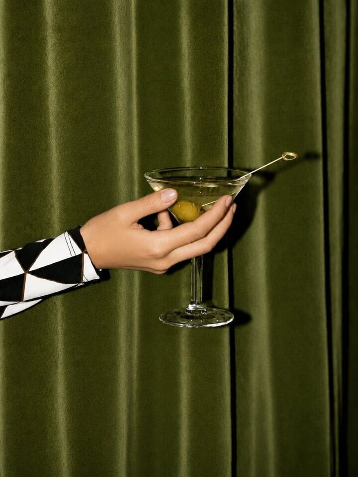 Taça de drink com azeitona contra cortina verde, à noite no Emma