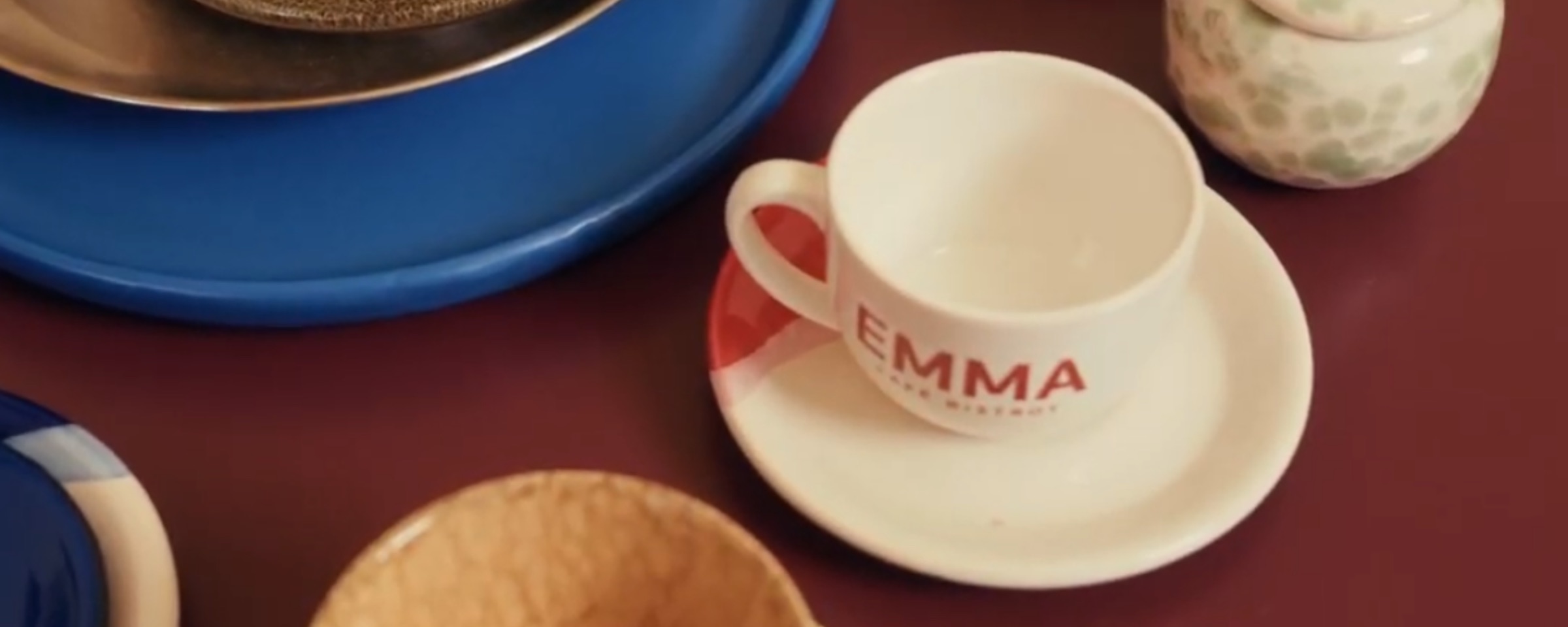 Xícara Emma Café Bistrot sobre mesa bordô, ao lado de louças e objetos de garimpo